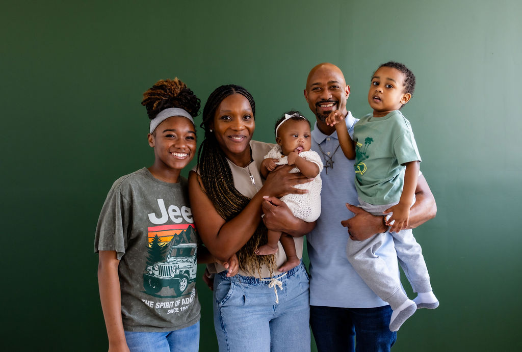 A family with parents, an infant, and older children posing against a green background